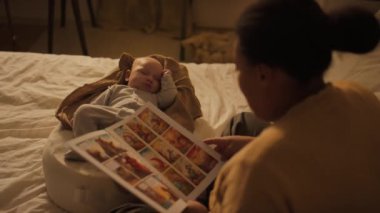 Siyahi annenin, 3 aylık erkek çocuğunun yuvada oturup renkli dinozor ailesinin maceralarını anlatan çizgi romandan yüksek sesle masal okuduğu orta omuzlu bir fotoğraf.