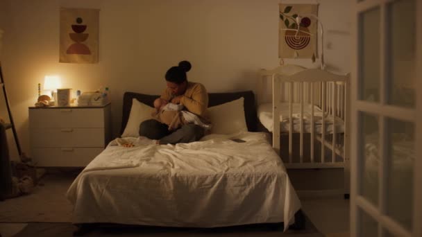 若いヒスパニック系またはアフリカ系アメリカ人の母親のワイドショットは 夕方に薄暗い寝室でベッドの上に足を横切って座って 彼女の新生児の娘を授乳します — ストック動画