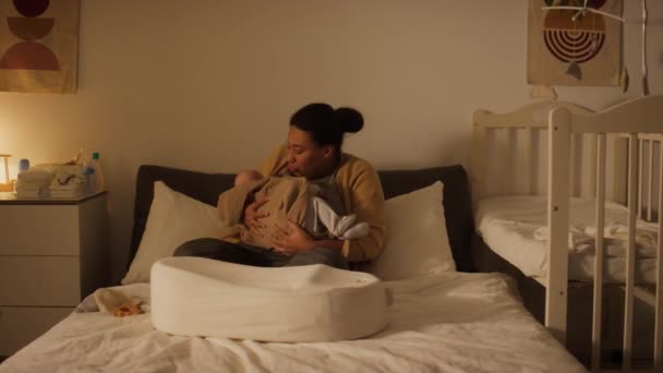 夜間に自宅でベッドに座っている若いアフリカ系アメリカ人の母親のフルショット 新生児の母乳育児 そして慎重に愛情のある視線でコクーンで眠るために彼を置く — ストック動画