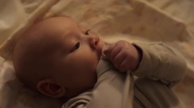 3 aylık erkek bebeğin, yatak çarşafında sırt üstü yatıp etrafa bakınırken, kollarını sallarken ve diş çıkarırken kollarını çiğnerken yakın plan çekimleri.