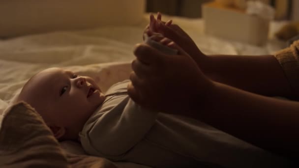 ベッドの上で横たわっているボディスーツの3ヶ月の男の子の幸せな笑顔のミディアムショット カメラを見て 彼の腕 脚と赤ちゃんの体操をしている認識できない母親の手 — ストック動画