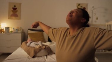 İspanyol ya da Afrikalı Amerikalı annenin yatak odasındaki fitness topunun üzerinde otururken, hamilelikten sonra kollarını kaldırırken ve zıplarken ve bebek koza içinde yatarken orta eğik pozu.