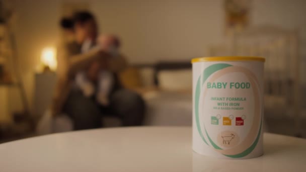 在家中卧房桌上放着婴儿奶粉罐的特写镜头 在模糊的背景下 无法辨认的母亲抱着和亲吻新生儿 — 图库视频影像