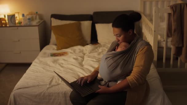 赤ちゃんの息子や娘が自宅でベッドに座ってスリングラップを貼り付け ラップトップを入力してレポートを書き 出産休暇中にパートタイムで働いている若い多民族の母親のミディアムショット — ストック動画
