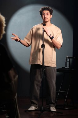 Genç komedyen ya da şovmen seyircilerin önünde duruyor ve stand-up performansı sırasında mikrofonla konuşuyor.