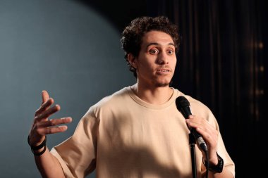 Stand-up kulübünün genç erkek komedyeni performans sırasında monoloğu telaffuz ederken mikrofonla konuşuyor ve el kol hareketleri yapıyor.