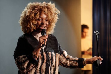 Happy young actress or comedian speaking in microphone while standing on stage in front of audience and performing new monologue clipart