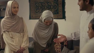 İki Müslüman kadının tesettüre bürünmüş ve abayas giyip bayram kutlamalarından önce evde genç bir adam tarafından takdim edilen anonim erkek misafirlerin önünde eğildiği orta ölçekli görüntüler.
