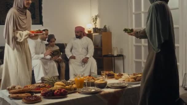 在开斋节上 两个年轻穆斯林用传统菜肴盖住阿巴约妇女餐桌的侧影 而家里的男人则在沙发上看古兰经 — 图库视频影像