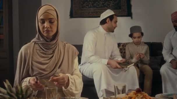 在Uraza Bayram与家人一起摆设节日餐桌 男人在沙发上讨论可兰经时 用中型手提式镜头拍摄年轻女子戴头巾放置餐具和向女性亲属进餐的场景 — 图库视频影像