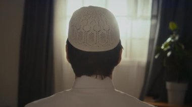 Arka sandıkta, beyaz kafatası şapkalı, isimsiz bir müslüman el kaldırıyor ve evde takbirlere dua ederken ezan okuyor.