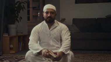 Beyaz başlıklı ve kandoralı sakallı bir müslümanın orta boy portresi evde oturmuş, elinde küçük kutsal Kur 'an kitabıyla kameraya bakıyor.