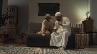Beyaz kandora 'da Müslüman bir babanın Kur' an okuyup kurallarını okuduğu ve Tasbih 'i tutarken oğlunun akşamları oturma odasında boncuk kullanmasını öğrettiği tam bir görüntü.