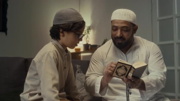 自宅で夕方に座ってコーランを唱える父親の後に繰り返し祈るための注意深い少年学習のフレーズのメディア映像 — ストック動画