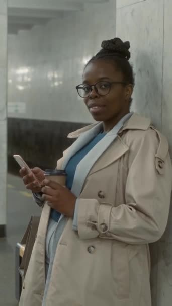 アフリカ系アメリカ人の女性が 現代地下鉄の地下鉄でスマートフォンとコーヒーカップを保持している彼女の地下鉄列車を待っている間 カメラと笑顔を見ながら眼鏡で描いた縦横の肖像画 — ストック動画
