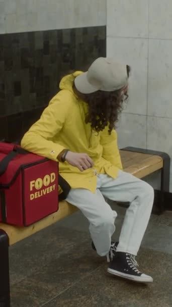 白种人卷发送货员身穿黄色夹克坐在长椅上 头戴大红包 在现代地铁里等火车的垂直全景画面 — 图库视频影像