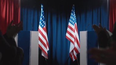 Kamu binasında tribünler, Amerikan bayrakları, mikrofonlar, iki olgun beyaz erkek ve kadın adayın yürüdüğü, el salladığı ve herkesin önünde alkışladığı orta boy bir sahne.