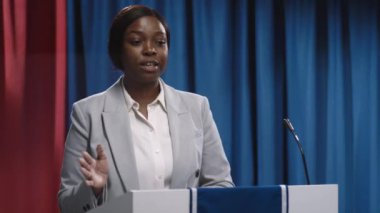 Kürsüde kürsünün arkasında durup seçim kampanyası sırasında siyasi gündem üzerine konuşma yapan gri takım elbiseli, kendinden emin genç Afrikalı Amerikalı kadın adayın orta boy yakın çekimi.