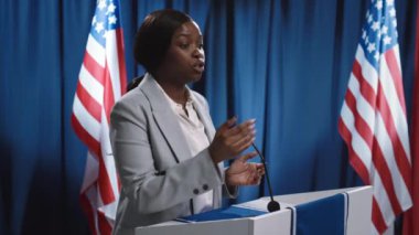 Resmi takım elbiseli genç Afrikalı Amerikalı bir kadının ABD bayrakları önünde kürsüye çıkıp güçlü siyasi açıklamalarda bulunurken, rakiplerini dinlerken, tartışırken ve aksini söylerken orta boy fotoğrafı.