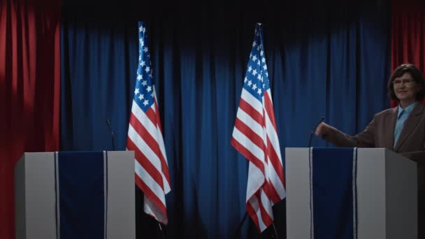 中全镜头在公共大厅的舞台上 两个玫瑰 美国国旗和麦克风 和中年白人男女政治家走在一起挥手问候选民 — 图库视频影像