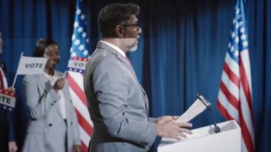 Orta yaşlı Hintli Amerikalı erkek başkanlık adayının orta yaşlı görüntüleri. Cumhuriyetçi partiden vali ya da başkan adayı. Kürsüye doğru yürüyorlar ve salondaki kalabalığı selamlıyorlar.