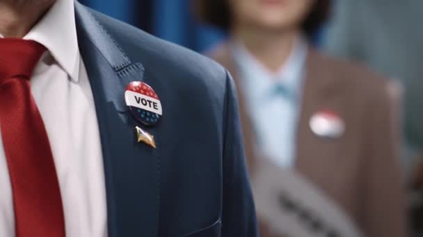 スーツ 白いシャツ 赤いネクタイ 投票と米国の旗バッジで認識できない男性候補者の上半身のクローズアップショットは 選挙大会中に心臓でスピーチしました — ストック動画