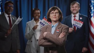 Beyaz kadın başkan, vali, senato ya da kongre adayının gülümseyen orta boy portresi, kollarını kavuşturmuş, gülümseyen, çok ırklı kampanya ekibiyle ABD bayrağı önünde poz veriyor.