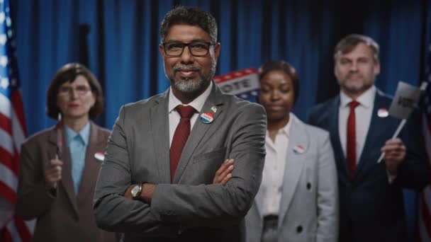 カリスマ的なインド系アメリカ人の男性大統領候補と彼の多様なチームが折りたたみ腕で立って 選挙運動中にアメリカの旗の前で微笑んでいる中立の肖像画 — ストック動画