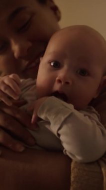 Orta boy dikey çekimde İspanyol anne kucağında 4 aylık bir bebek, sarılıyor ve gülümsüyor, çocuk parmağını çiğniyor ve kameraya bakıyor.