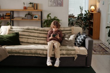 Evdeki geniş oturma odasında rahat koltukta oturan sevimli küçük çocuk akıllı telefon ekranında online video izliyor.