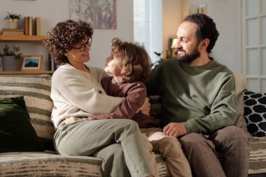 Oturma odasında kameranın önünde otururken gülümseyerek birbirlerine bakan mutlu, genç bir aile.
