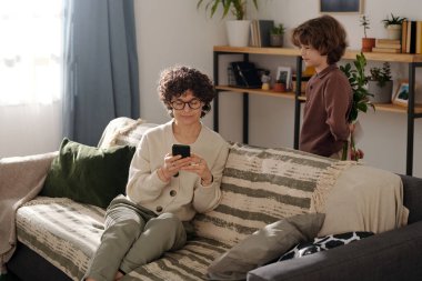 Sevimli bir çocuk, annesinin önünde kanepede akıllı telefon kullanırken arkasında bir demet çiçek ve el yapımı kartpostal saklıyor.