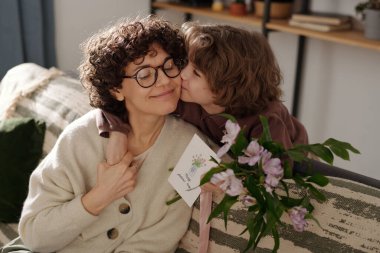 Sevimli çocuk annesini yanağından öpüyor ve kanepeye yaslanıp onu kucaklıyor ve bahar tatilinde tebrik ediyor.