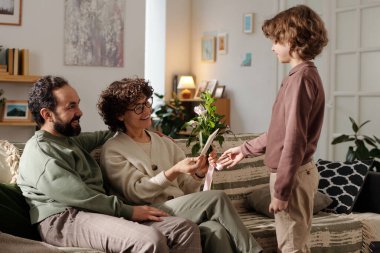 Kanepede oturan ve önünde duran tatlı oğlunun el yapımı kartpostalında selamlarını okuyan mutlu genç bir kadın.