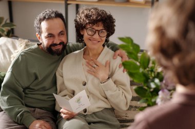 Mutlu genç bir adam güzel karısını kucaklarken ikisi de oğlunun önünde kanepede oturup annesi için çiçek topluyor.