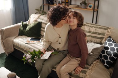 Sevecen bir anne bir demet çiçekle ve kartpostalla sevimli oğlunu kucaklıyor ve kanepesinde otururken onu yanağından öpüyor.