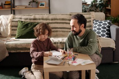 Küçük çocuk babasının yanına oturup Anneler Günü için tebrik kartı hazırlarken pastel boyalarla bir şeyler çiziyor.