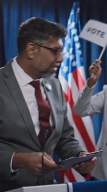 Orta yaşlı, kendine güvenen orta yaşlı Asya kökenli Amerikalı erkek başkan adayının sahnede yürüdüğü, gülümsediği, seyirciyi selamladığı, el salladığı, seçim kampanyası için düzenlediği basın toplantısında orta boy dikey çekimi.