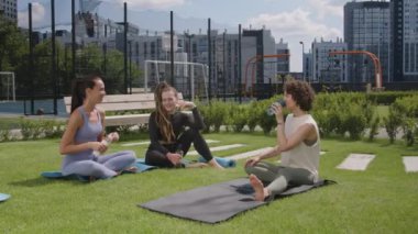 Aktif giyimli üç beyaz kadının şehir parkında egzersiz minderlerinde otururken ve antrenmandan sonra dinlenirken geniş görüntüler.