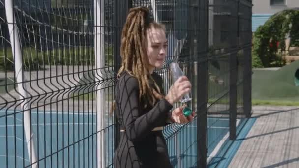 在阳光灿烂的日子里 白种人漂亮的女运动员靠在娱乐区的栅栏上 训练后喝着可怕的水 — 图库视频影像