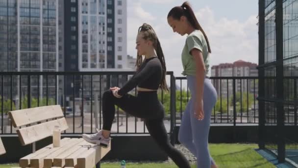 素晴らしい夏の日に屋上レクリエーションエリアでトレーニングする前に 怪我防止のために足を伸ばすスポーツウェアの2人の白人運動女性のサイドフル映像 — ストック動画