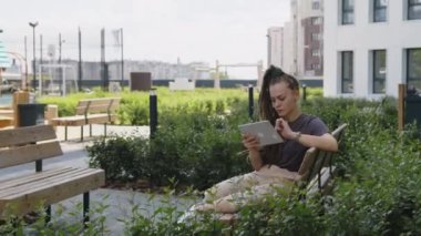 Kafkasyalı iş kadını Rastalı ve günlük kıyafetleriyle tablet bilgisayarda çalışıyor boş bir çatı parkındaki ahşap bankta oturuyor.