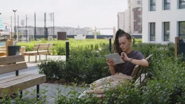旁边的全景画面是白种人的女商人 带着可怕的头发 穿着休闲装 在平板电脑上远程工作 坐在空荡荡的公寓屋顶公园的木制长椅上 — 图库视频影像