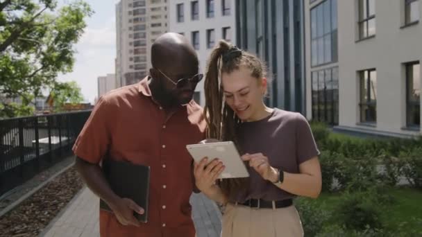 都市公園の散歩中に陽気なアフリカ系アメリカ人のアソシエイトにタブレット上のプロジェクトのアイデアを提示するドレッドロックを持つ白人ビジネスマンのメディア映像 — ストック動画