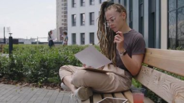 Modern parktaki ahşap bankta oturan ve dijital tabletteki verilerle eşleşen evrakları kontrol eden gözlüklü, düşünceli öğrenci ya da iş kadınının yan görüntüleri.