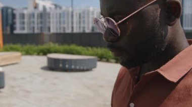Sıcak yaz günlerinde modern çatı parkında güneş gözlükleriyle oturan, dizüstü bilgisayarda kod yazan siyahi erkek BT çalışanının yan çekimleri.