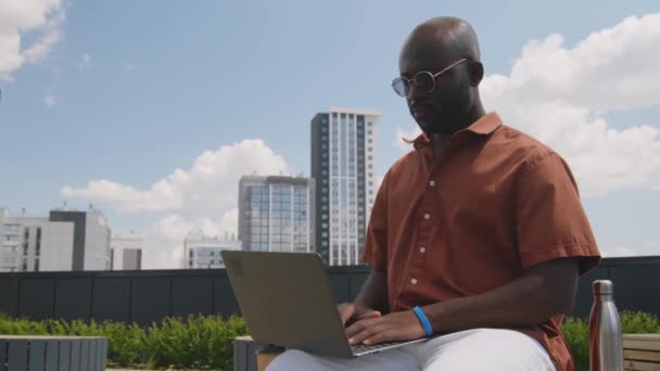 サングラスで深刻なアフリカ系アメリカ人のフリーランサーのメディア映像 晴れた夏の日の都市の屋根公園でラップトップコンピュータでオンラインで作業 — ストック動画