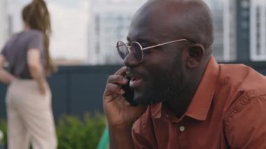 Yazın çatıdaki parkta otururken cep telefonuyla konuşan, altın güneş gözlüklü Afro-Amerikan sakallı adamın yakın çekimi.