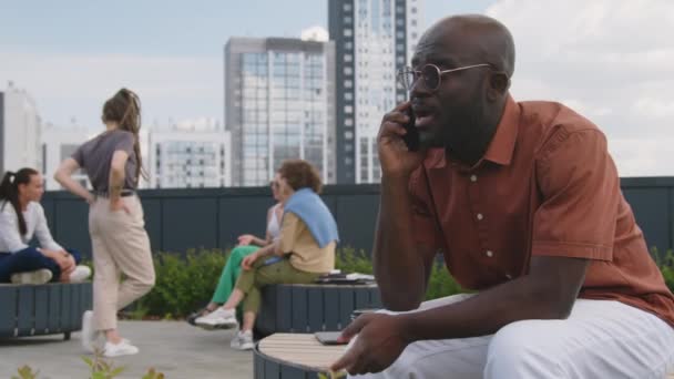 テラコッタのシャツとサングラスの不満の黒人のビジネスマンの側面映像は 夏の日に現代の屋上公園での仕事の問題について緊張して電話で話します — ストック動画
