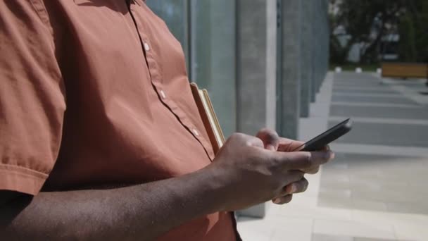 テラコッタのシャツで認識できないビジネスマンのサイドミッドセクション映像は オフィスビルや携帯電話でのメッセージに傾いています — ストック動画